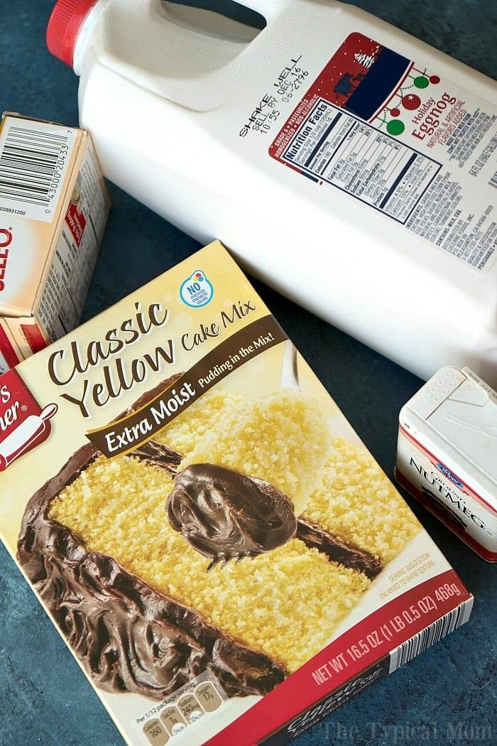 A box of classic yellow cake mix sits on a blue surface, accompanied by a carton of milk, and unsalted butter. The cake mix box showcases an image reminiscent of an egg nog pudding cake adorned with chocolate frosting.