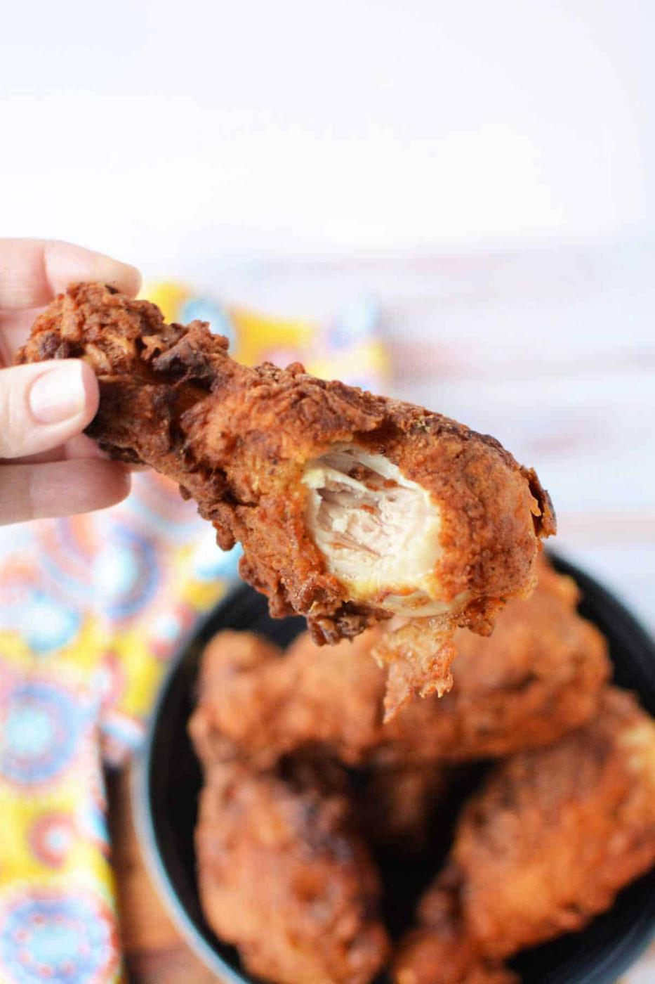 A person holds a piece of fried chicken with a bite taken, hovering it over a bowl filled with more crispy delights, all resting on a vibrant cloth. The golden hue suggests an expertly prepared egg wash recipe that adds to the tantalizing crunch and flavor.