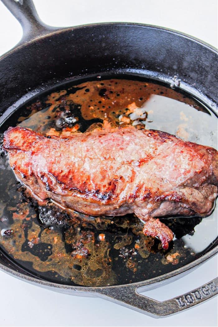 A juicy flank steak sizzles in a cast iron skillet against a pristine white background.