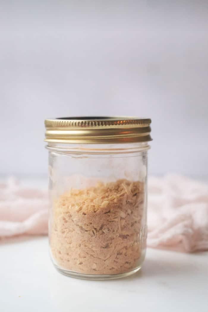 A clear jar with a gold lid, filled with homemade French Onion Soup Mix, rests elegantly on a white surface.