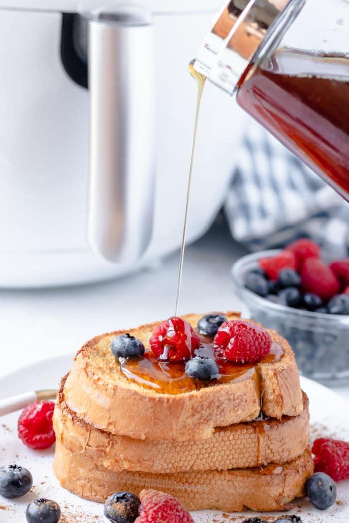 Stack of French toast, perfectly crisped in the air fryer, topped with berries and drizzled with syrup, with a bowl of berries in the background.