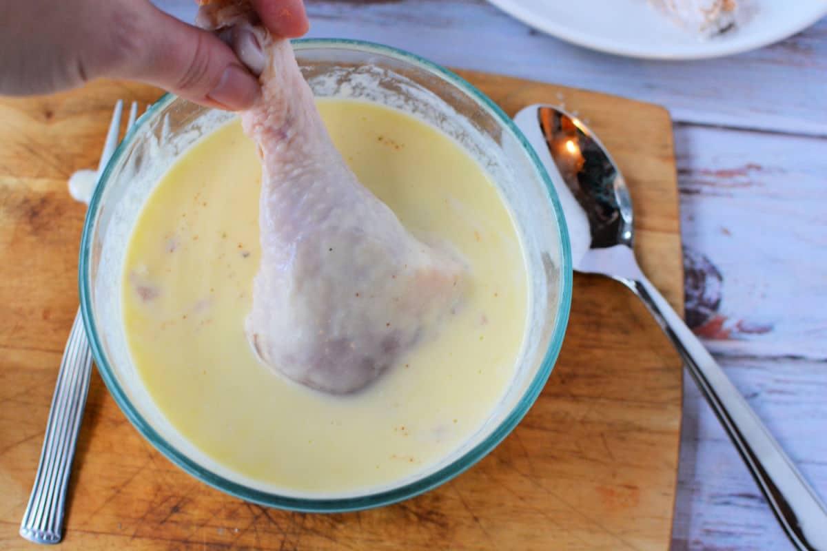 Hand dipping raw chicken legs into creamy batter in a glass bowl on a wooden board, ready to be transformed into delicious pan fried chicken legs, with a spoon and fork beside it.