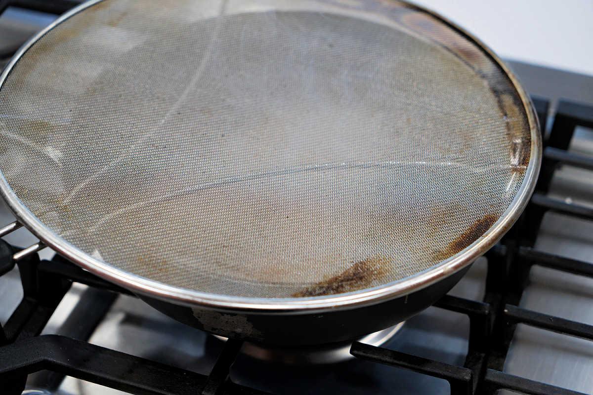 A frying pan on a stove, covered with a metal mesh splatter screen, is perfect for preparing your favorite fried tilapia recipe.