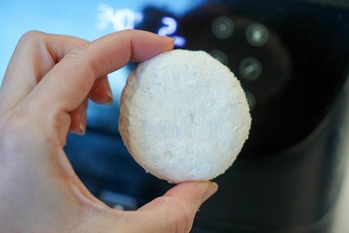 A hand holding a round, powdery white biscuit—perfectly cooked like frozen biscuits in air fryer—in front of a blurred digital control panel.