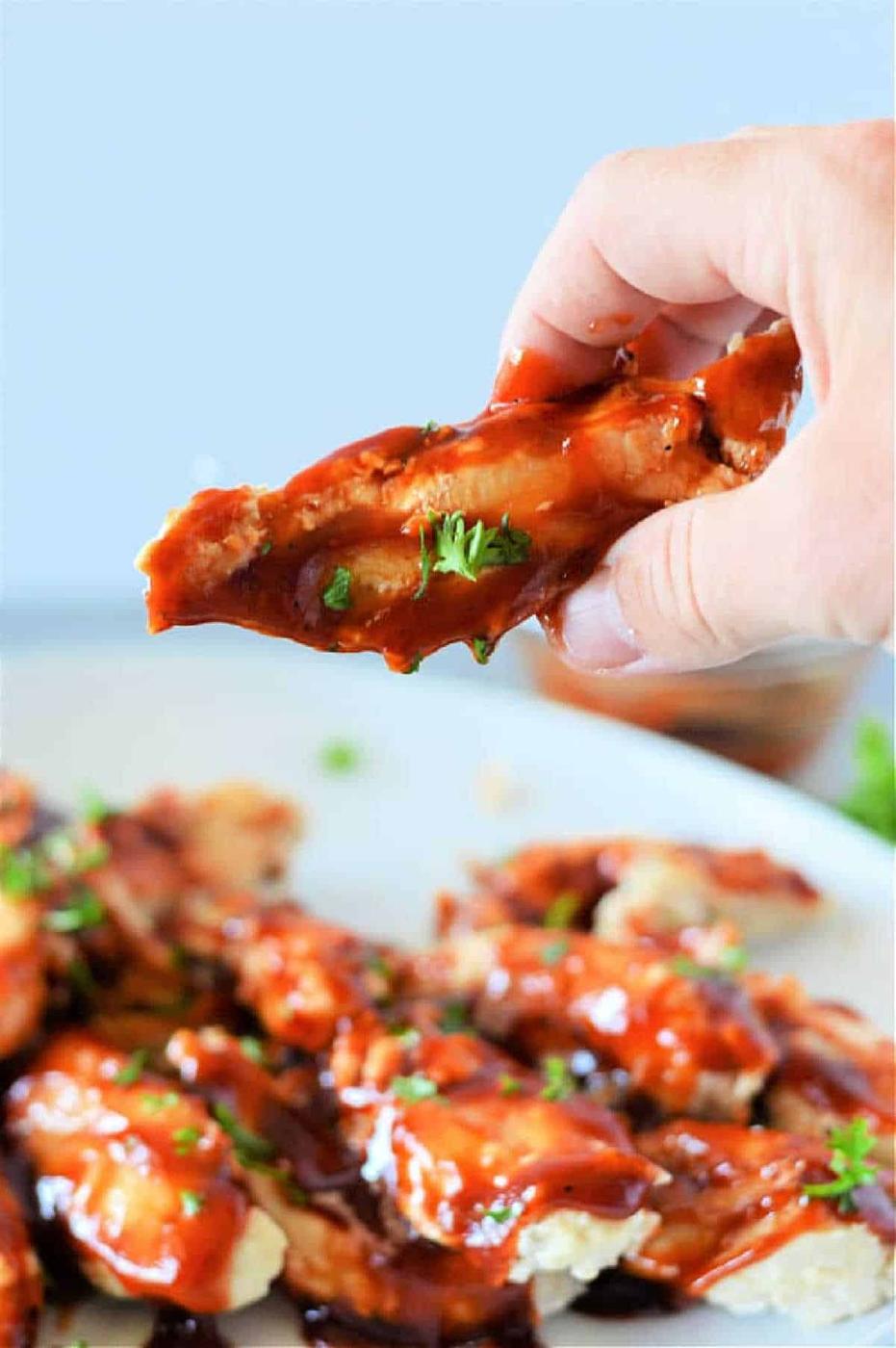 A hand holds a saucy chicken wing above a plate of barbecue wings, garnished with chopped herbs—perfect inspiration for making frozen chicken tenders in the Instant Pot at home.
