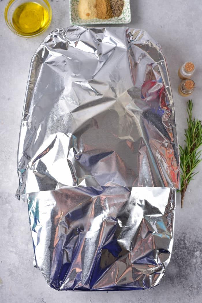 A frozen duck inside a baking dish, which is covered with aluminum foil and centered on the marble countertop. Nearby are a small bowl of olive oil, a plate with seasoning, two small shakers, and a sprig of rosemary.