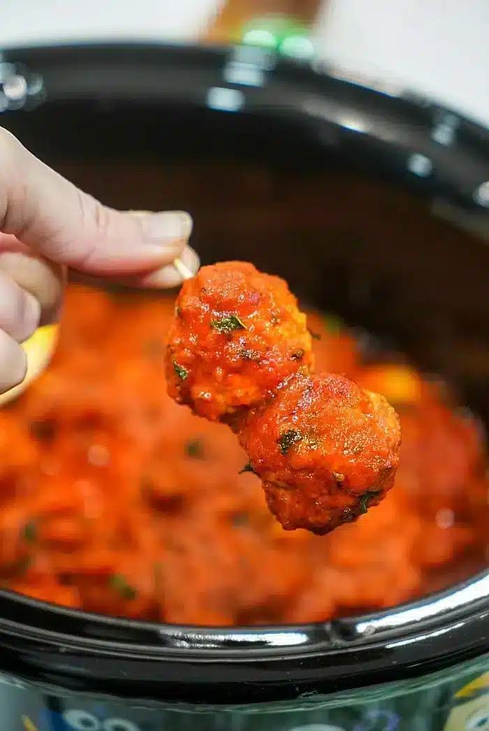 A hand holds a skewer with two succulent meatballs coated in tomato sauce, poised above a crockpot filled with more simmering meatballs and sauce. Using frozen meatballs in the crockpot makes this dish an effortless delight.
