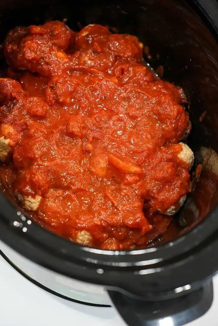Frozen meatballs in crockpot simmer, covered in rich tomato sauce inside a black slow cooker.