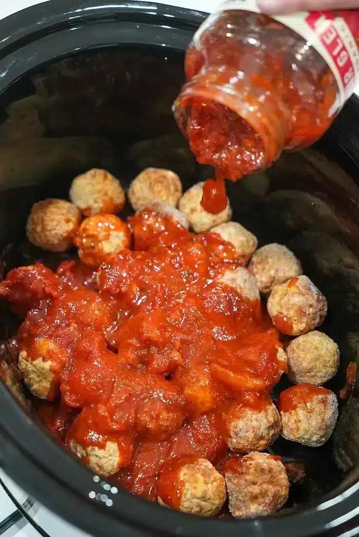 Frozen meatballs in the crockpot are being covered with tomato sauce poured from a jar.