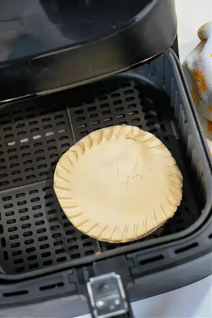 A frozen pot pie with a crimped edge and cracked top sits in the basket of an air fryer. The interior surface, complete with black perforated grates, is visible. On the side, a partial view of a patterned cloth adds a touch of charm.