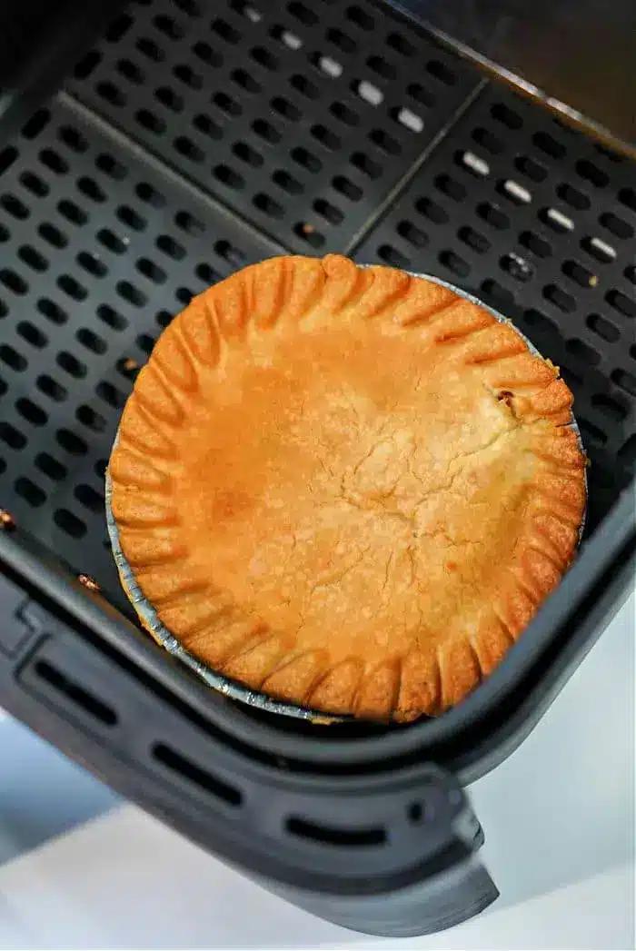 A golden-brown frozen pot pie sits in an air fryer basket, its crust evenly baked with a decorative crimped edge. The air fryer features a perforated base for optimal air circulation, making it perfect for transforming frozen delights into crispy perfection.