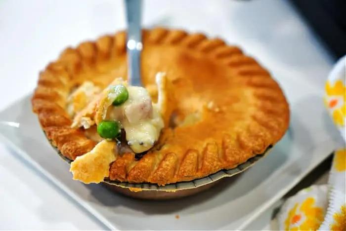 A fork lifts a bite of creamy chicken pot pie with peas and carrots, fresh from a frozen pot pie cooked to perfection in an air fryer. The flaky, golden-brown crust has a beautiful fluted edge, and a patterned cloth is partially visible beside the white plate.