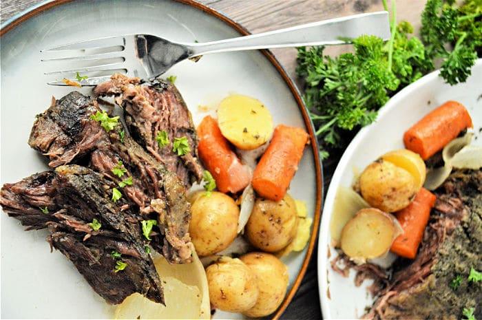 Plate of roast beef, carrots, and potatoes garnished with parsley, next to a serving dish of the same meal—perfect for preparing frozen roast in a slow cooker for tender, flavorful results.