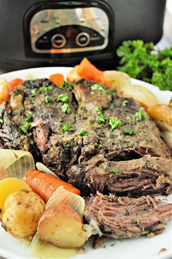 Tender pot roast with carrots, potatoes, and onions on a white plate in front of a slow cooker—perfect for making even a frozen roast in the slow cooker.