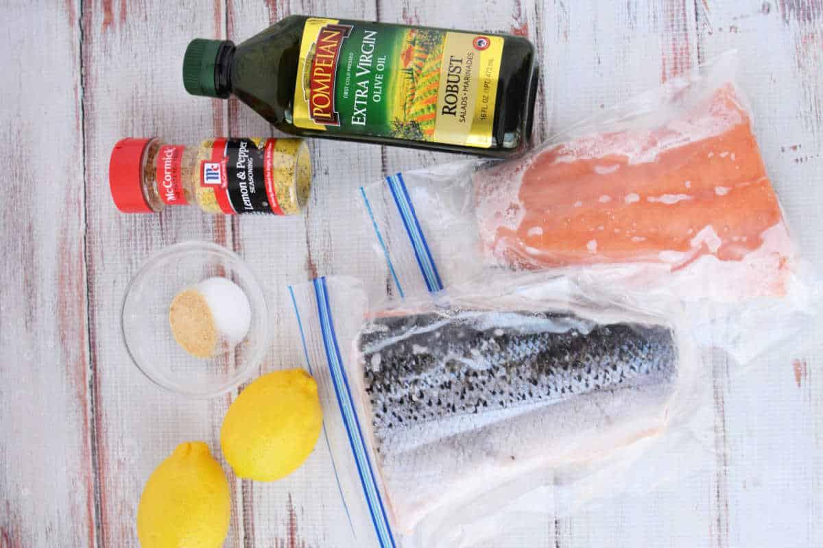 Two raw or frozen salmon fillets, olive oil, spices, lemons, and seasonings are arranged on a white wood surface, ready to be cooked in the oven.