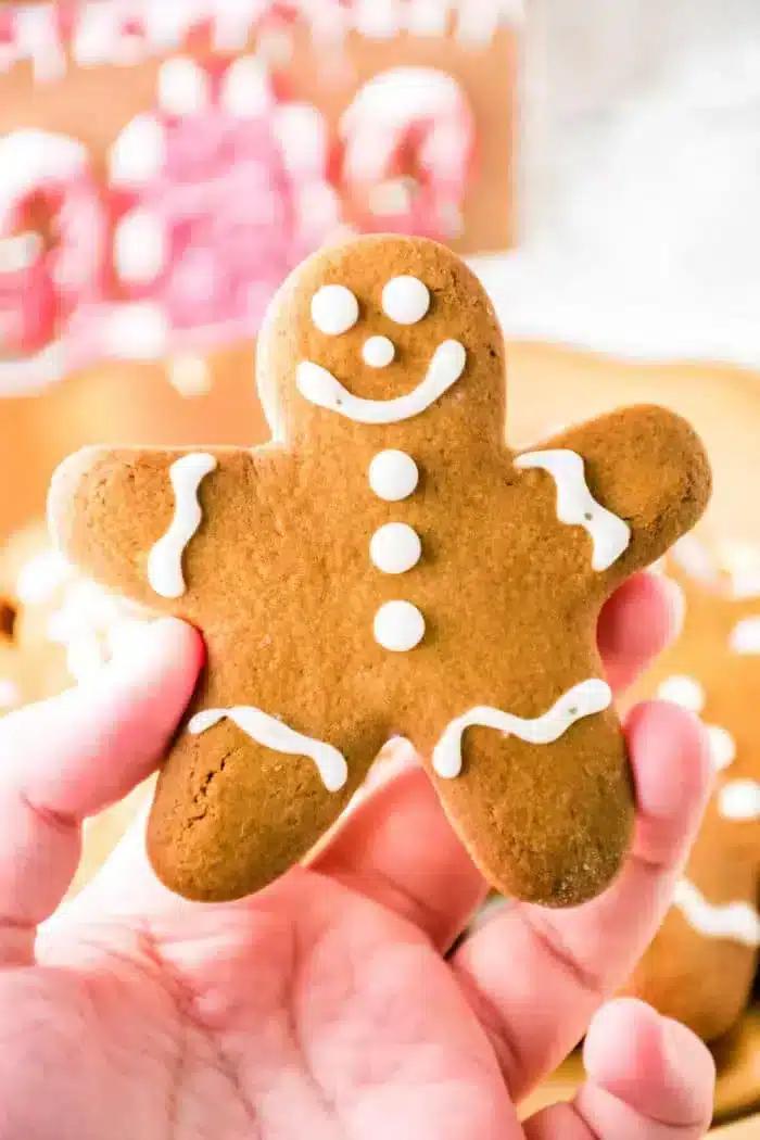 A hand holds a gingerbread cookie, crafted from spiced gingerbread dough, shaped like a person and adorned with white icing for eyes, mouth, buttons, and outlines. Other similar cookies softly blur into the background.