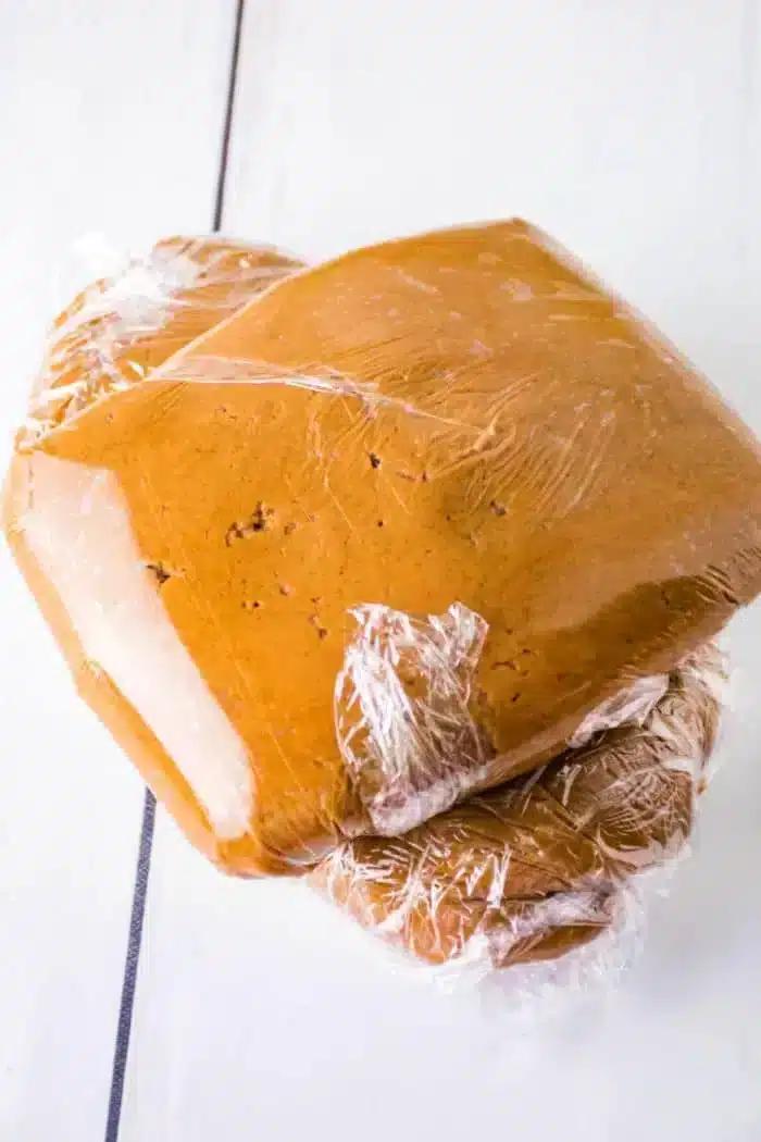 Three stacks of gingerbread dough wrapped in clear plastic wrap are placed on a white surface.