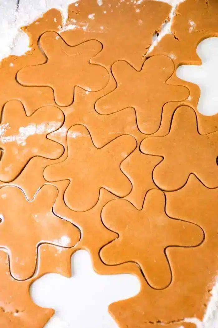 Close-up of rolled-out gingerbread dough with flower-shaped cutouts. The dough is light brown with a few flour patches on the surface. Some cutouts are fully formed, while others remain connected to the surrounding dough.