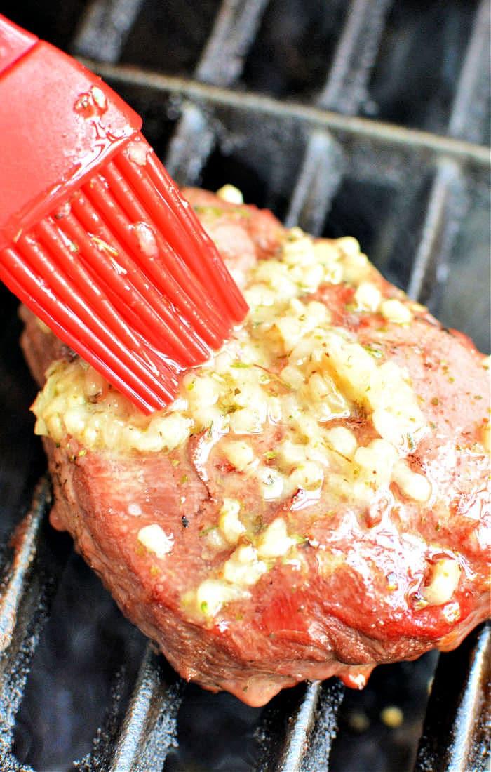 A red brush delicately applies garlic and herbs to a frozen steak on the grill, showcasing an essential step in the art of how to grill frozen steak to perfection.