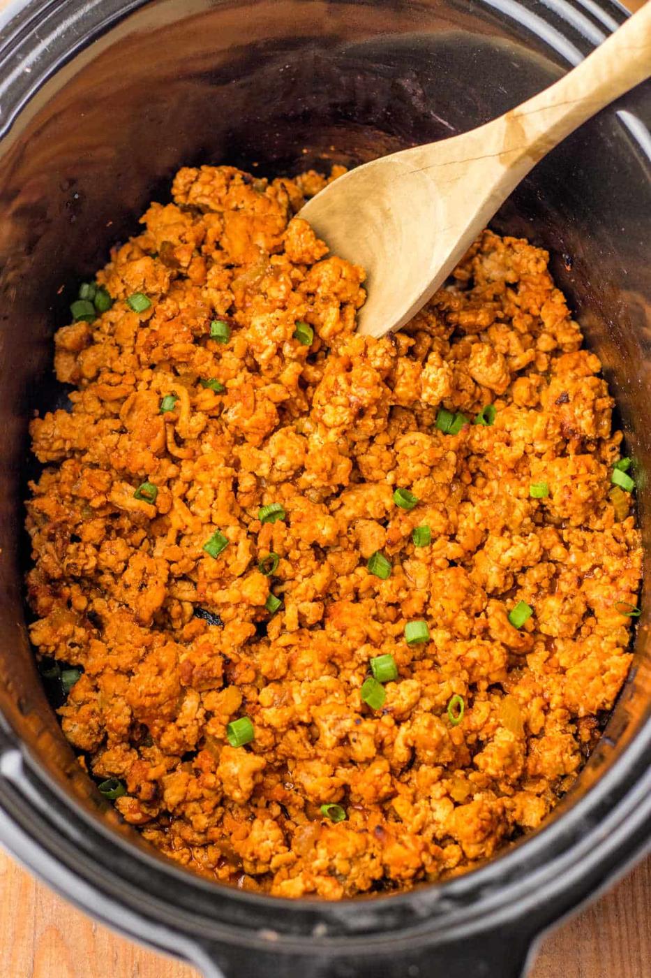 Cooked seasoned ground turkey in a crockpot with a wooden spoon, transforming it into a delightful dish reminiscent of Ground Chicken Sloppy Joes, garnished with chopped green onions.