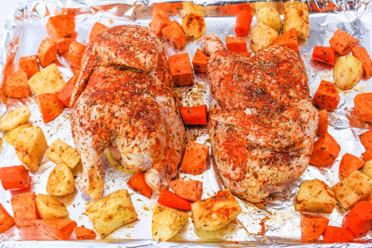 A seasoned half chicken rest amid chunks of sweet potatoes and carrots on a foil-lined baking sheet.
