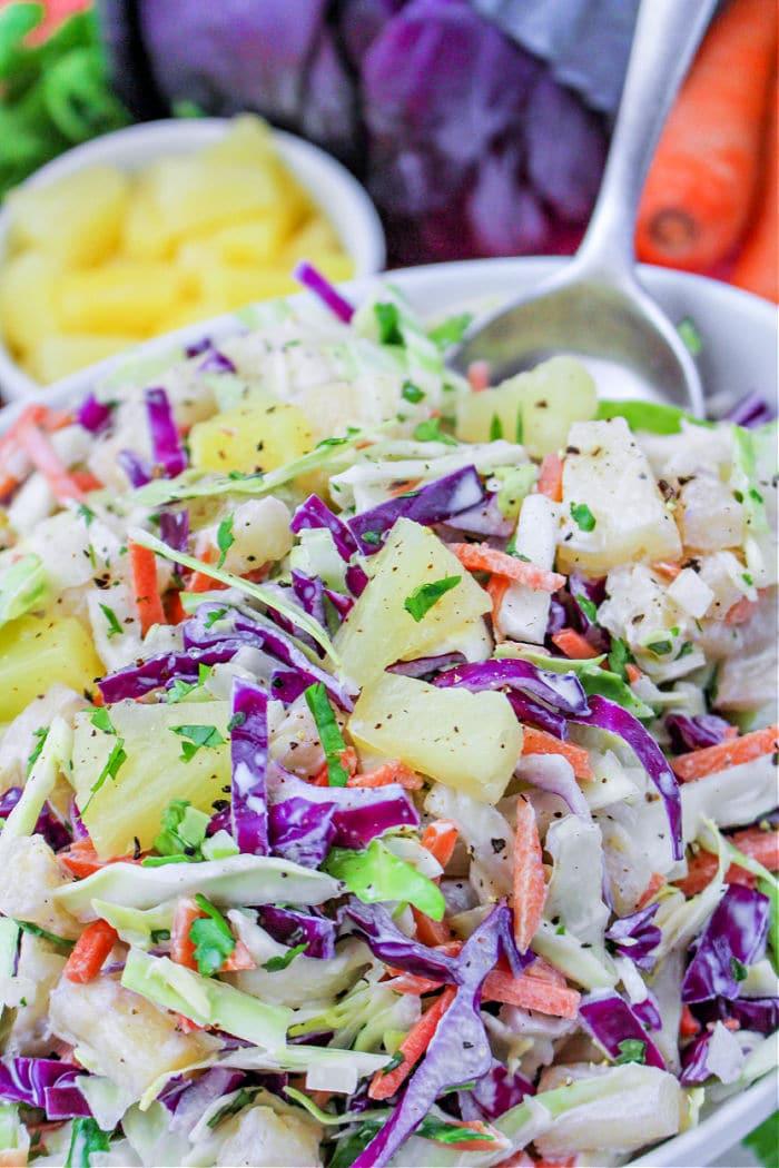 Hawaii coleslaw features colorful cabbage, carrots, and juicy pineapple chunks in a white bowl, sitting beside a spoon and fresh ingredients.