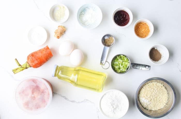 Various ingredients, including eggs, carrot, oil, seasonings, flour, and ground chicken for Homemade Chicken Nuggets with Ground Chicken, arranged on a white countertop.