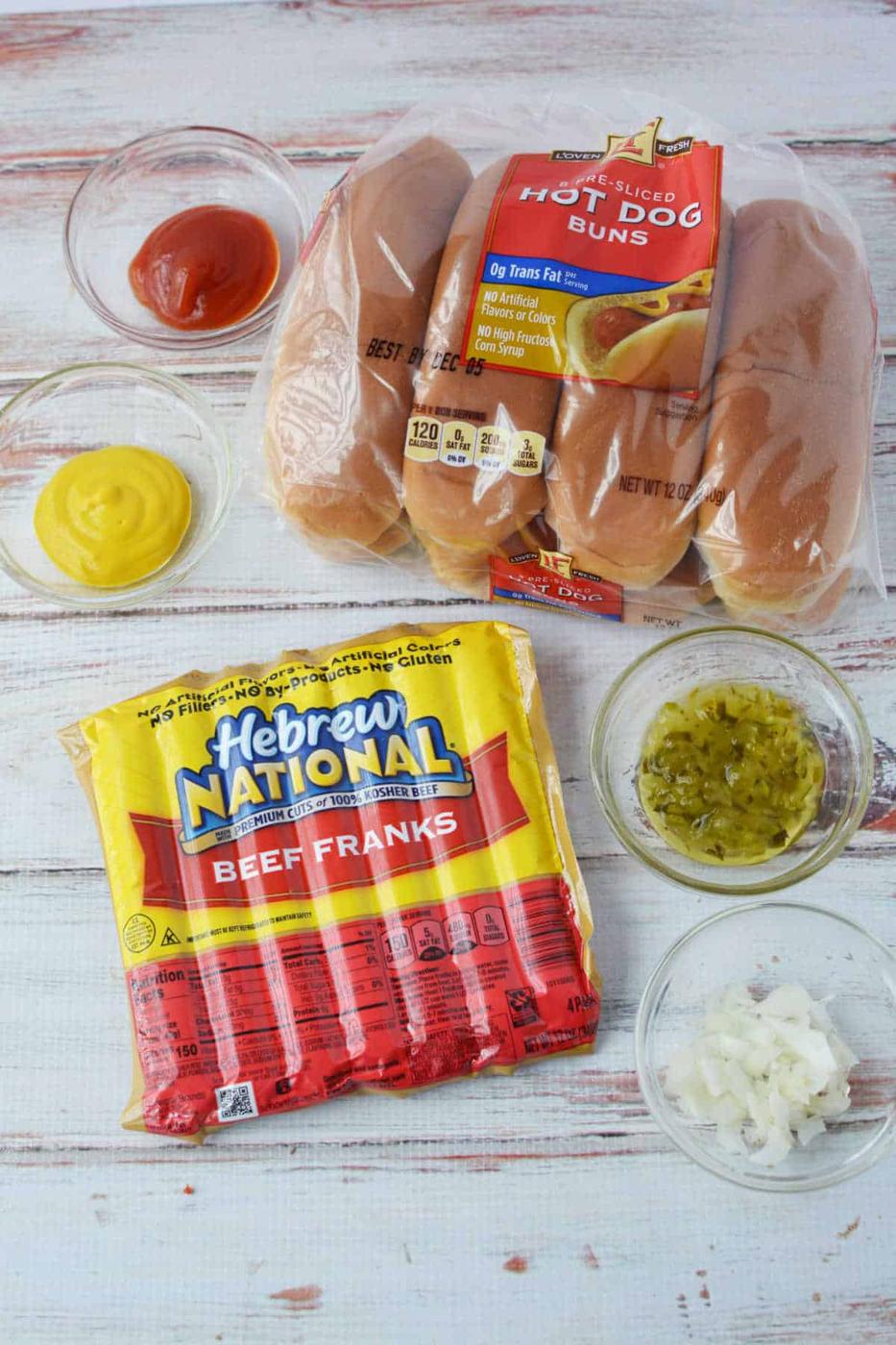 Baked Hot dog ingredients: buns, beef franks, ketchup, mustard, relish, and chopped onions rest on a wooden surface, ready to create perfect hot dogs in the oven.