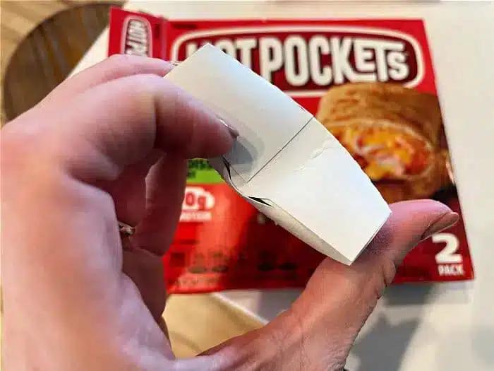 A hand holding an unfolded crisping sleeve with a Hot Pockets package in the background.