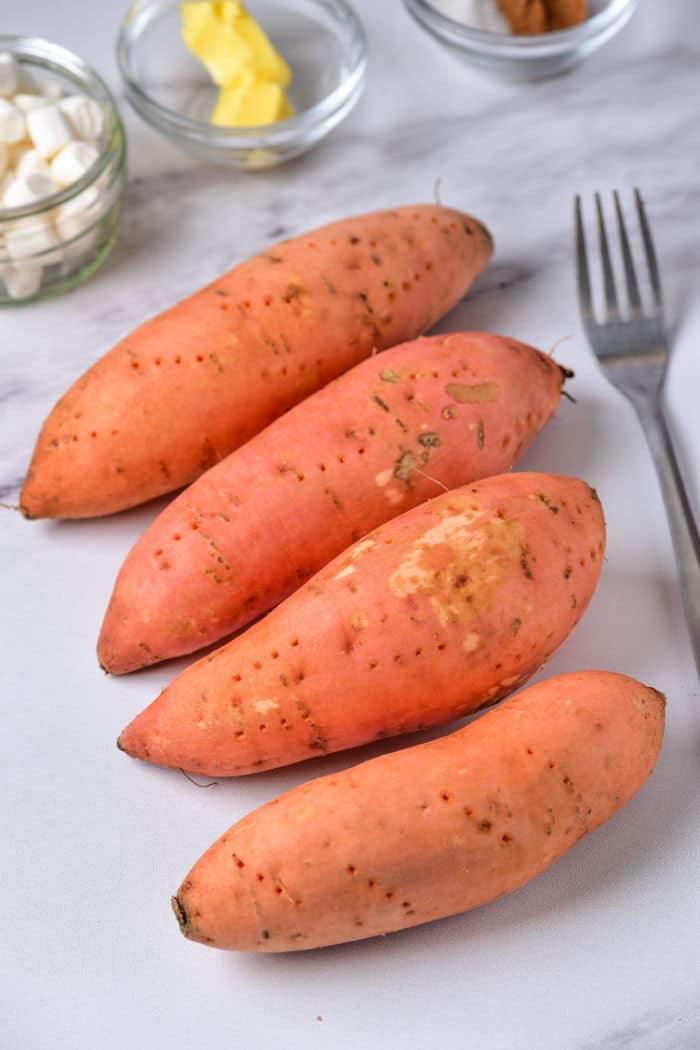 Four raw sweet potatoes rest on a white surface with a fork nearby, ready to reveal how long to bake a sweet potato at 400.
