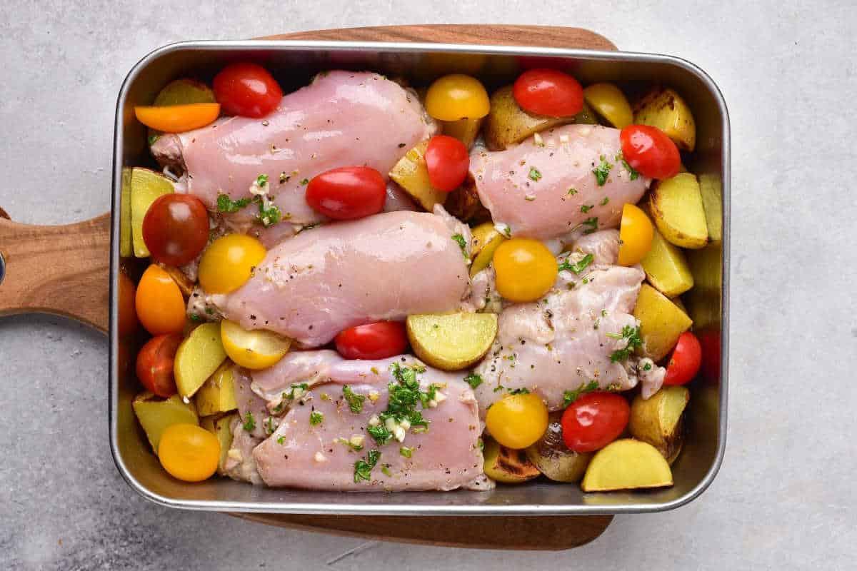 Raw chicken thighs, cherry tomatoes, and potato wedges in a baking pan, ready to bake at 375.