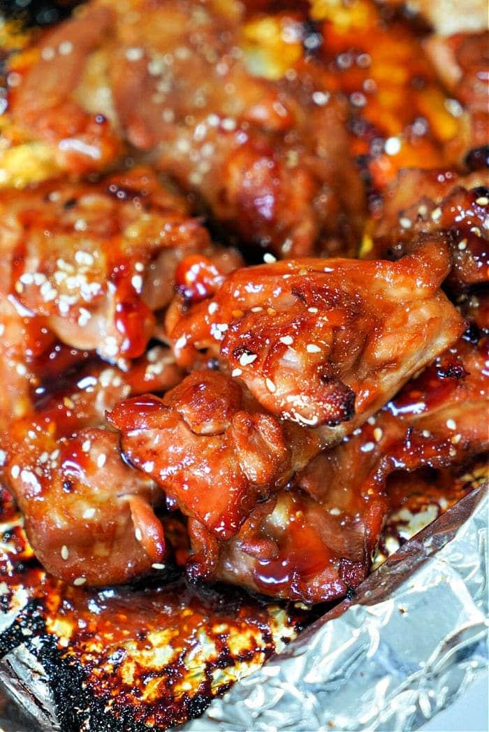 Close-up of glazed chicken thighs with sesame seeds on foil, showcasing a caramelized and sticky texture reminiscent of teriyaki chicken thighs.