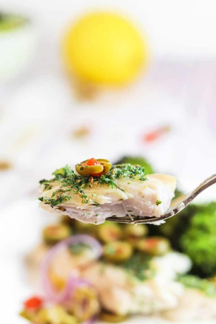 A fork holds a bite of fish topped with herbs and sliced olives, with blurred veggies in the background—a tempting scene for anyone curious about how long to bake mahi mahi to perfection.