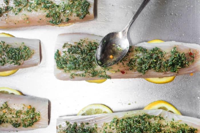Raw fish fillets on lemon slices with herbs being spread by a spoon, creating a perfect pairing while pondering how long to bake mahi mahi for that ideal, flaky texture.