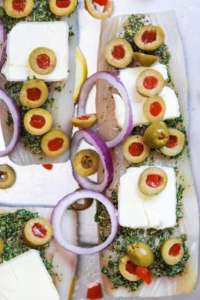 Raw fish fillets topped with herbs, butter, green olives, red onion rings, and pimentos on a tray—perfect for trying out how long to bake mahi mahi for tender, delicious results.