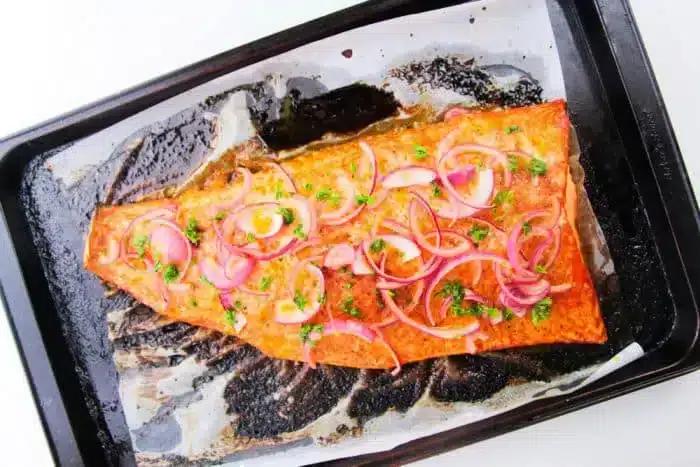 A cooked salmon fillet lies on a parchment-lined baking tray. The salmon is garnished with thinly sliced red onions and chopped parsley. Wondering how long to bake salmon at 400? The tray shows some residue from the cooking process, indicating it was baked to perfection.