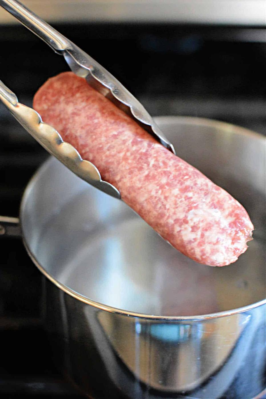 Tongs holding raw brats over a pot of boiling water on the stove, as you ponder how long to boil brats for that perfect flavor.