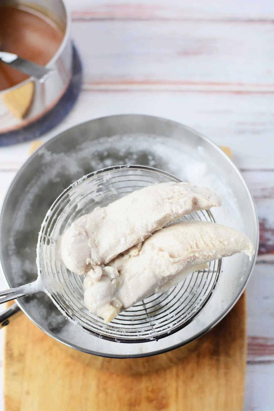 Two cooked chicken tenders, demonstrating how long to boil perfect tenders, are being lifted from a pot of boiling water with a wire skimmer and placed on a wooden board.