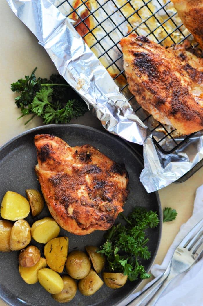 Baked chicken breast with roasted potatoes and parsley on a plate, next to more chicken on a foil-lined rack. Wondering how long to cook chicken breast in oven at 350? Its about 25-30 minutes for juicy perfection.