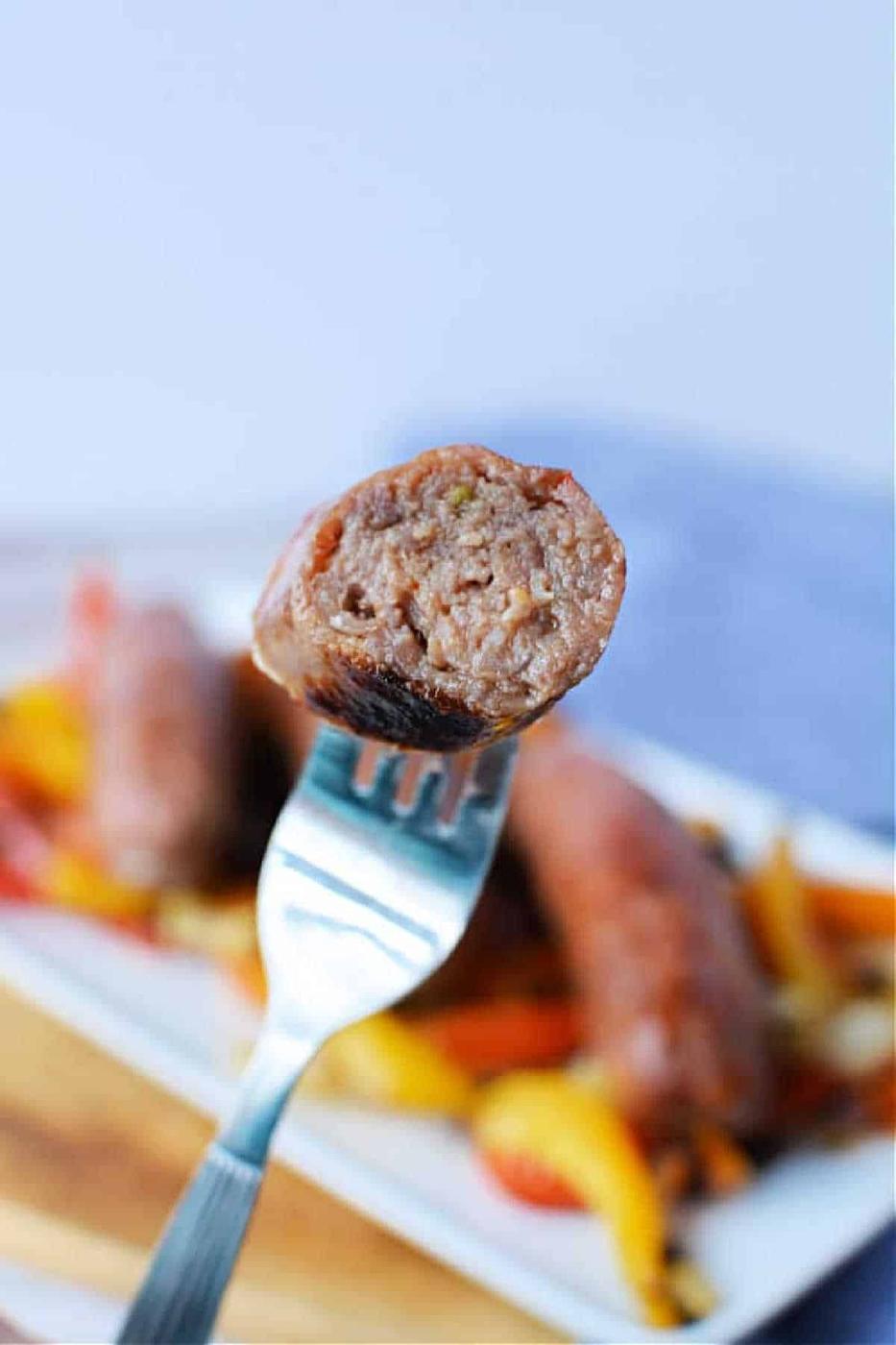 A fork holds a bite of cooked Italian Sausage in Oven, with peppers and onions blurred in the background.