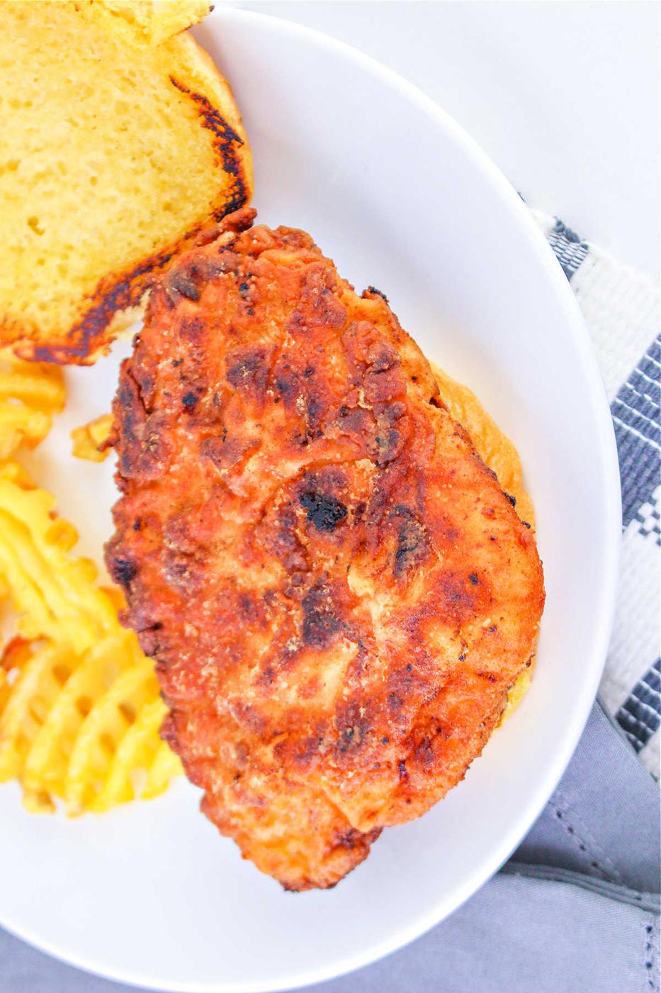 A fried chicken sandwich on a bun, perfectly crisp after learning how long to deep fry chicken breast, served with crinkle-cut fries on a white plate.