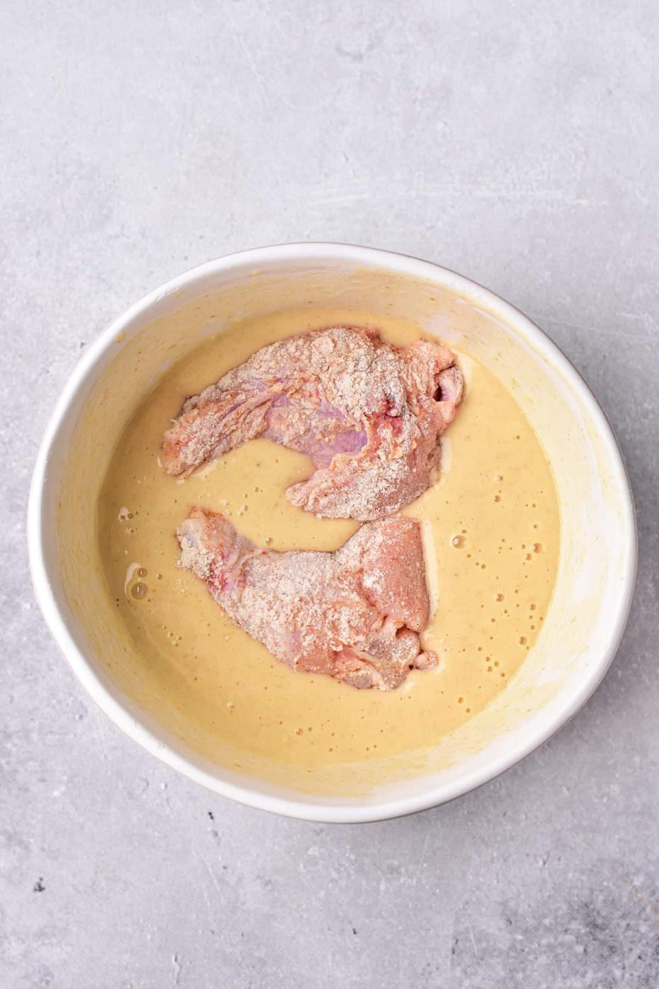 Raw chicken pieces in a creamy tempura batter sit invitingly in a round white bowl on a gray surface, while you ponder how long to deep fry chicken wings for that perfect crispiness.