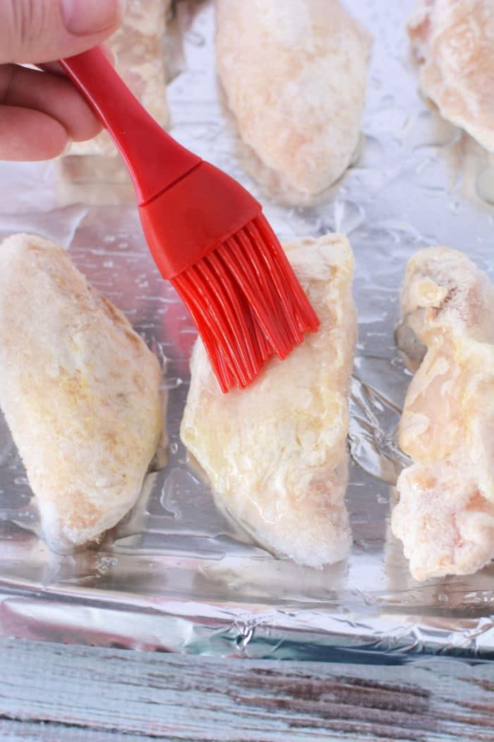 A hand demonstrates how to bake frozen chicken wings using a red basting brush to apply oil on them, neatly arranged on a foil-lined baking sheet.
