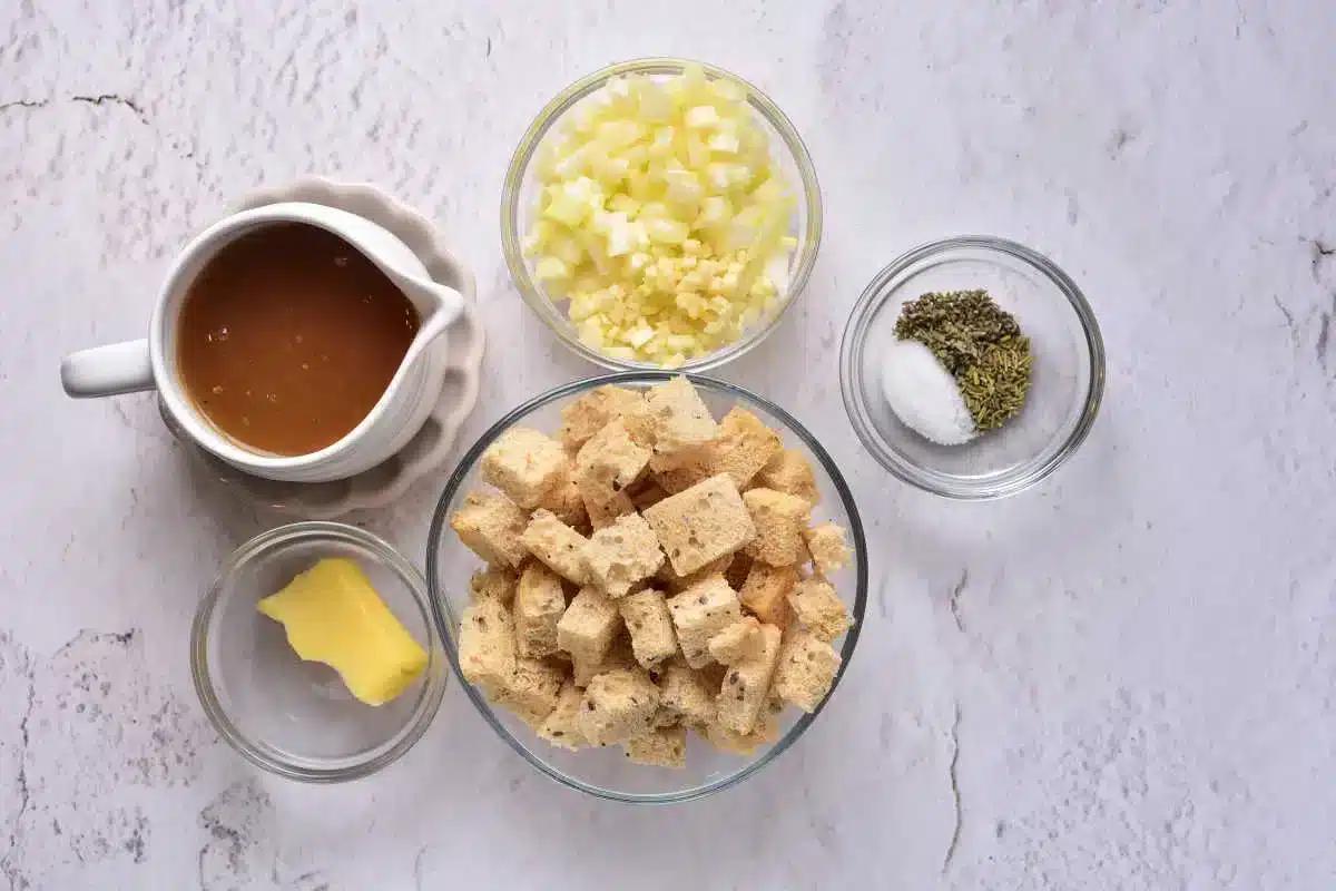 Ingredients for how to cook stuffing in the oven are laid out on a white textured surface: a pitcher of brown liquid, diced onions and garlic in a small bowl, dried herbs with salt in another, croutons in a medium bowl, and a pat of butter resting on a small plate.