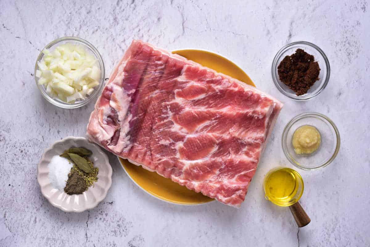 Raw pork belly rest on a yellow plate, surrounded by small bowls of spices, chopped onions, and oil on a marble surface—a perfect starter kit for those eager to learn how to confit pork.