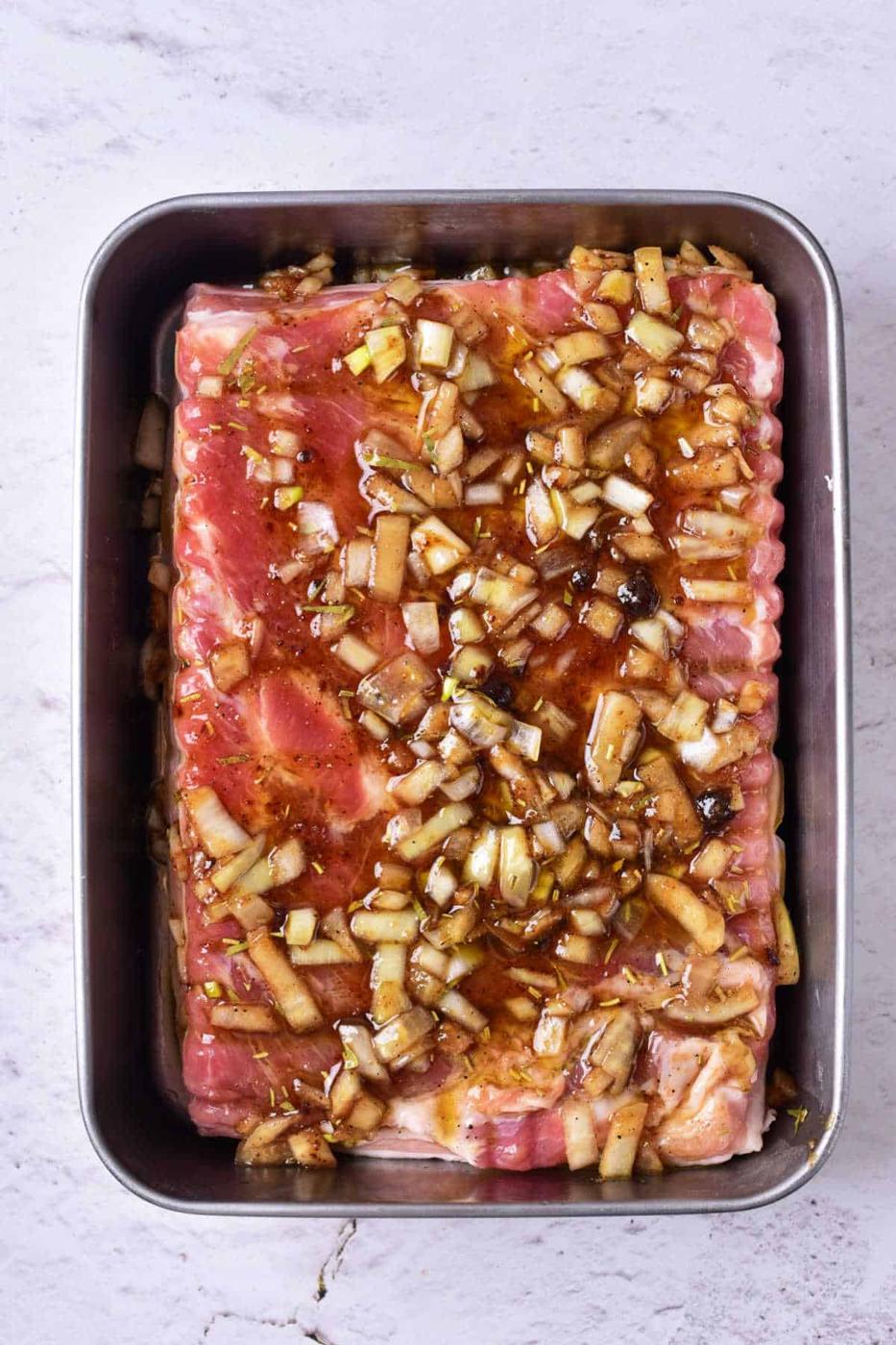 Raw pork belly marinated with chopped onions and sauce rest enticingly in a rectangular metal tray on a light gray surface, evoking the initial steps in learning how to confit pork with incredible flavor depth.