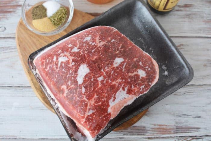 A block of frozen roast, sits on a black tray atop a wooden board. Next to it, a small glass bowl contains a mix of salt, pepper, garlic powder, and dried herbs. A bottle of Worcestershire sauce is partially visible in the corner.