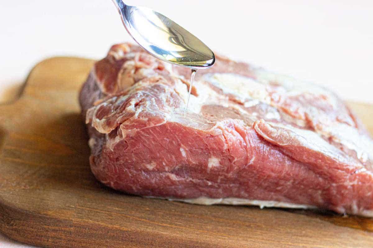 A spoon drizzling oil onto a raw piece of meat on a wooden cutting board sets the stage for mastering how to cook a New York Strip Roast.