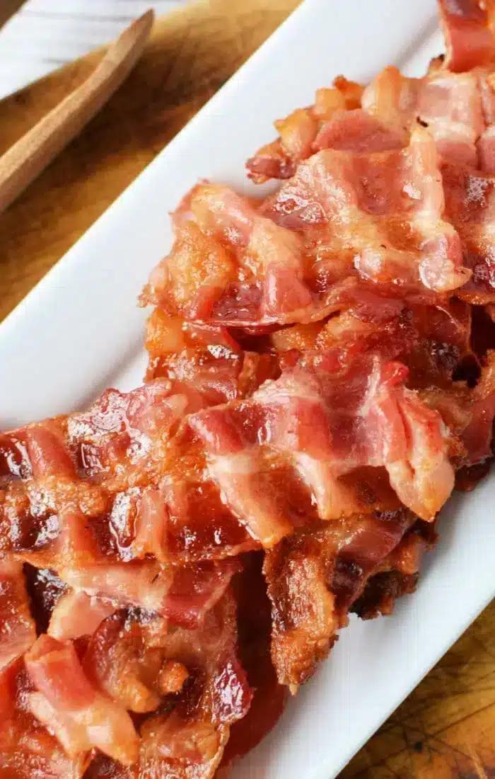 A plate of cooked, crispy bacon strips is arranged on a white rectangular platter. The bacon looks glossy and browned,