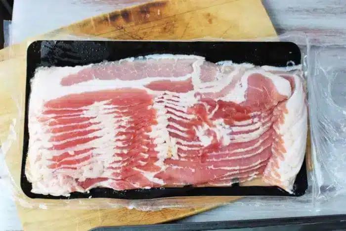 A sealed package of uncooked bacon strips is placed on a wooden cutting board.